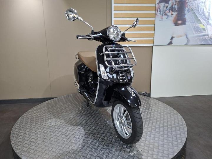 A black Vespa scooter with a tan seat displayed on a rotating platform in a showroom setting, with a light-colored wall and digital screen in the background.