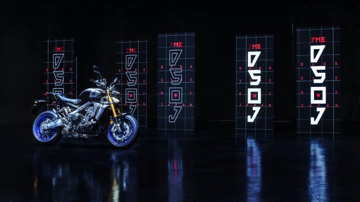 A modern motorcycle with metallic and black accents is positioned on a reflective surface, surrounded by tall digital displays featuring glowing red and blue text that reads "THE DARK SIDE OF JAPAN." The background is dimly lit, enhancing the vibrant colors of the motorcycle and screens.