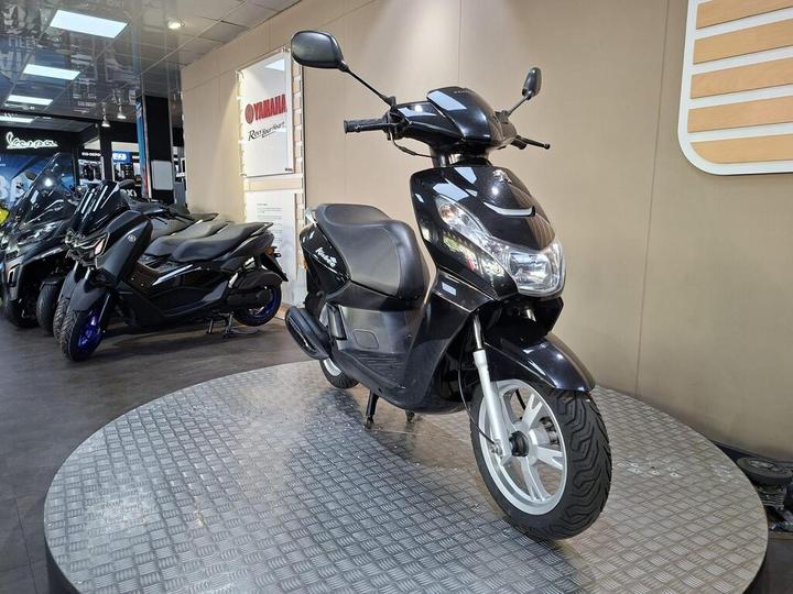 A black scooter is displayed on a circular platform in a showroom, with other scooters in the background. The wall behind features a Yamaha logo and promotional materials.