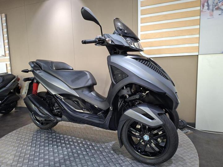 A modern gray scooter displayed in a showroom setting, featuring a sleek design with a black seat, red suspension spring, and black wheels.