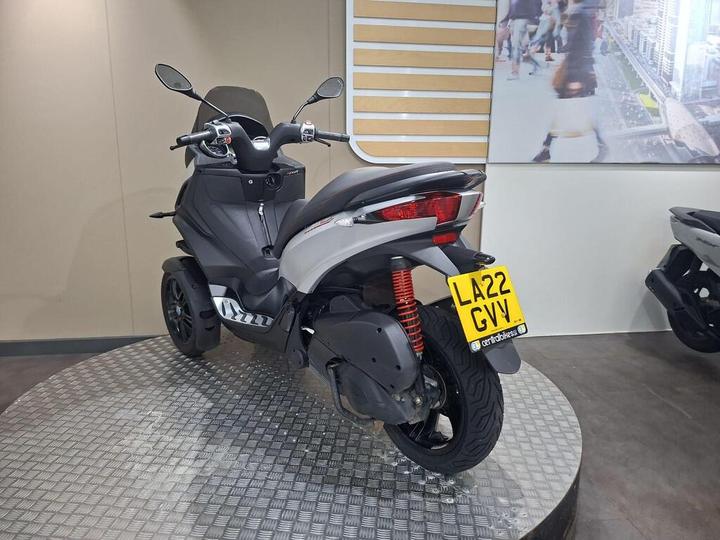 A sleek, modern three-wheeled scooter is displayed on a checkered platform in a showroom, featuring a gray body and black accents. The vehicle has a license plate reading "LA22 GVY" and is positioned against a light-colored wall with a cityscape graphic.