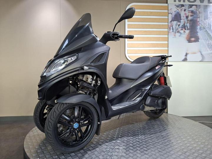 A sleek black three-wheeled scooter is displayed on a textured platform with a light-colored wall in the background and an abstract image of people shopping.