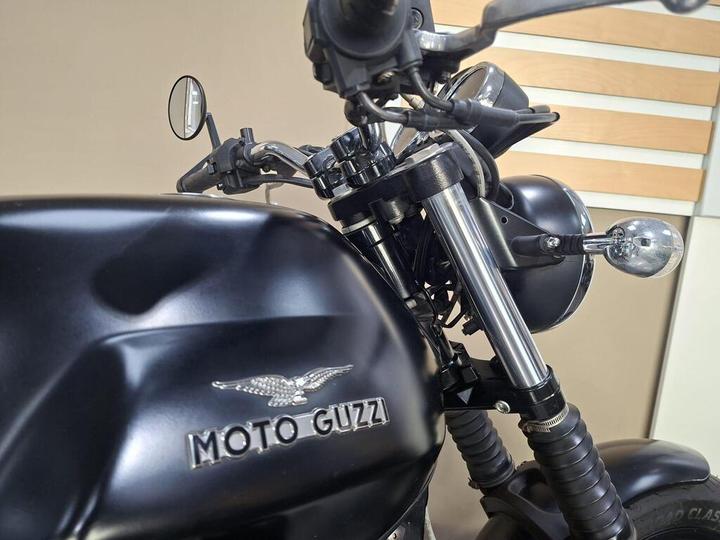 A close-up view of a black Moto Guzzi motorcycle, highlighting the handlebars, mirrors, and emblem on the fuel tank, set against a neutral background.