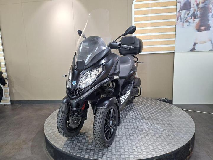 A black three-wheeled scooter displayed on a circular platform with a textured surface, featuring a windshield and storage compartment. The background includes a light-colored wall with a large photo of a crowd.