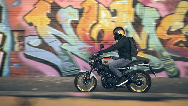 A rider in a black jacket and helmet rides a Yamaha XSR125 motorcycle past a colorful graffiti wall, capturing motion with a blurred background.