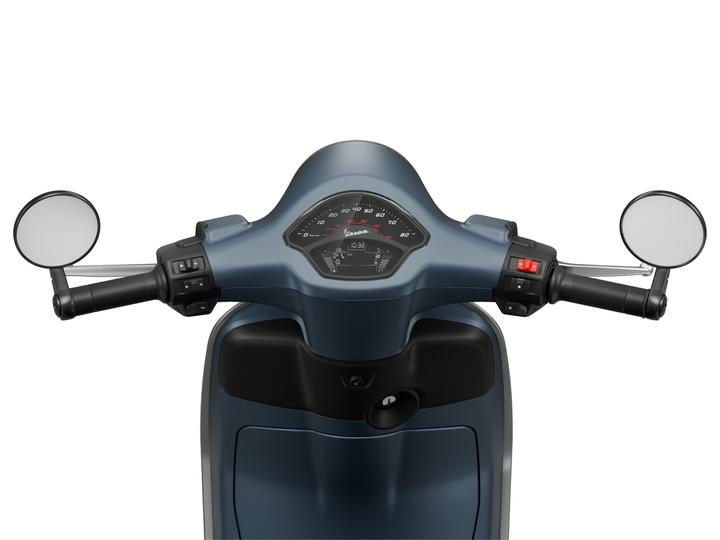 Close-up view of a scooter's handlebars featuring a black grip on either side, control switches on the left, a speedometer and dashboard in the center, and mirrors extending on both sides. The scooter has a sleek blue design.