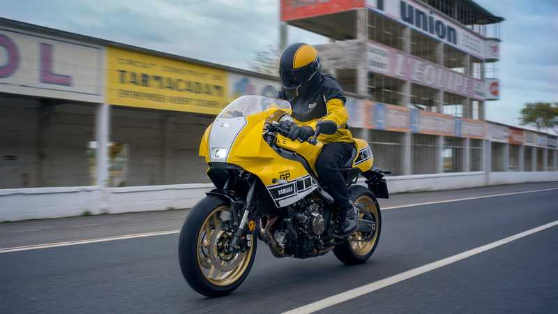 A motorcyclist in a yellow helmet and jacket rides a yellow Yamaha motorcycle on a racetrack, with blurred background signage indicating speed and motion.