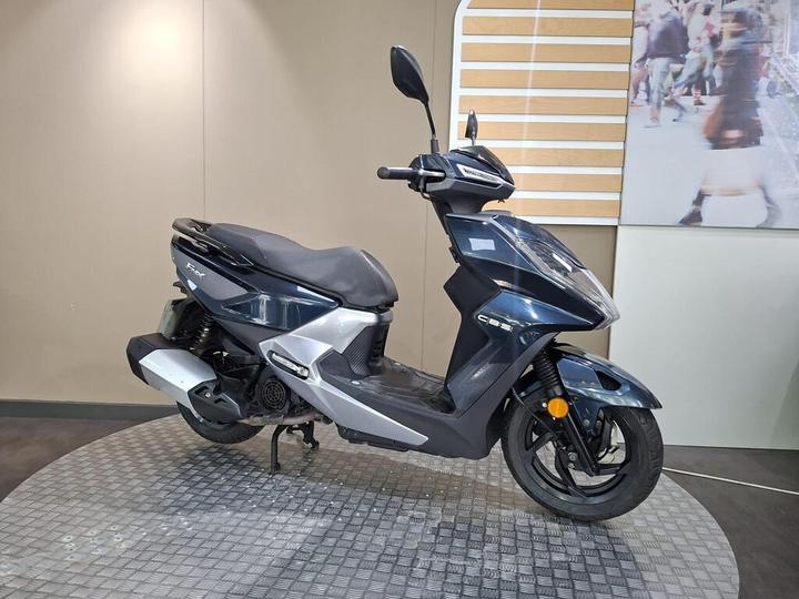 A blue and silver scooter is displayed on a circular patterned platform against a neutral background.