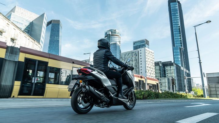 A motorcyclist wearing a black helmet and jacket rides a sleek scooter on an urban road, with modern skyscrapers and a tram visible in the background under a clear blue sky.