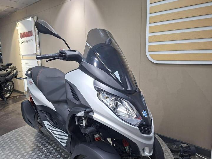 A close-up view of a Yamaha scooter in a showroom, showcasing its sleek design, rounded windshield, and ergonomic features, with a blurred background of other motorcycles.