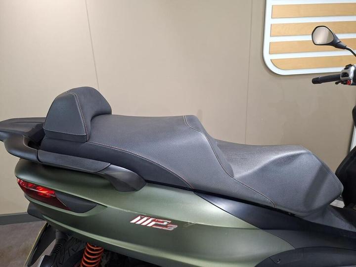 A close-up view of a motorcycle seat, featuring a black textured surface with red stitching, mounted on a green scooter body with visible suspension components. The background is a neutral-colored wall with wooden accents.
