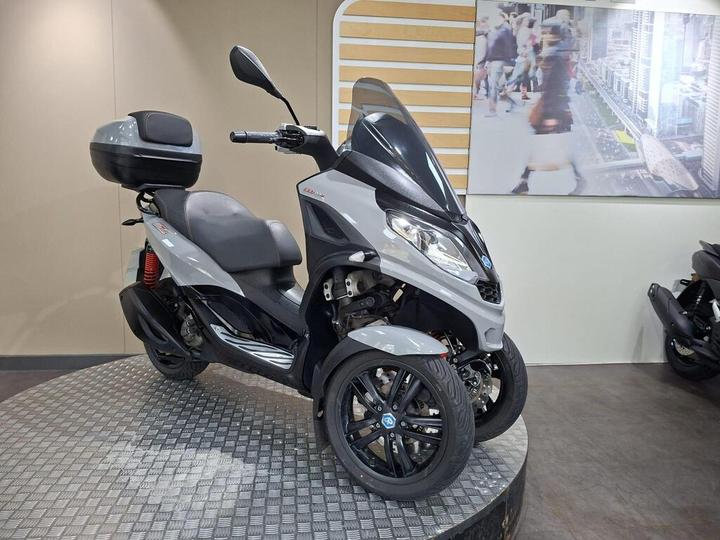 A gray three-wheeled scooter is displayed on a circular platform, featuring a black storage box on the back and a modern design. A blurred urban scene is visible in the background.
