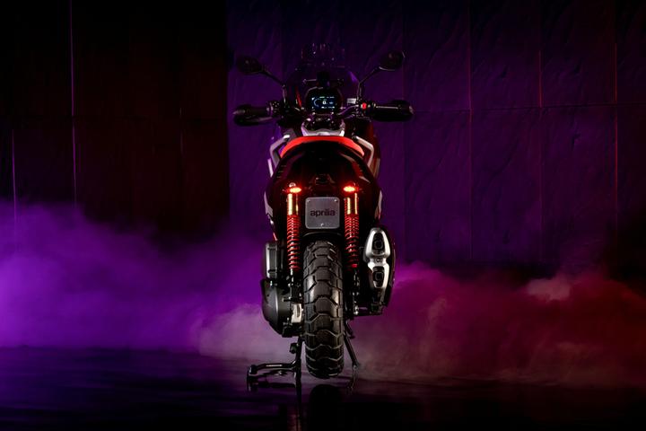 A rear view of a motorcycle, illuminated with purple and pink lighting, showcasing its sleek design, including a prominent tail with red lights and a patterned rear tire. Smoke adds a dramatic effect to the scene.