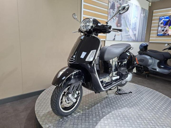 A sleek black scooter displayed on a circular metallic platform in a modern showroom, with another scooter partially visible in the background.