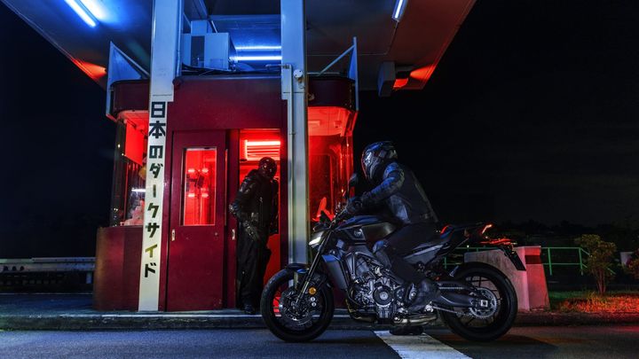 A motorcyclist in all-black gear sits on a sleek motorcycle near a dimly lit toll booth, with a figure in a black suit and helmet standing in the entrance, illuminated by red and blue lights at night. Japanese characters on the booth read "Japan's Dark Side."