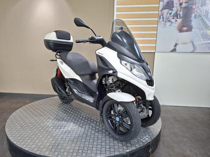 A sleek, white and black three-wheeled scooter displayed on a circular metal platform, showcasing its modern design and features.