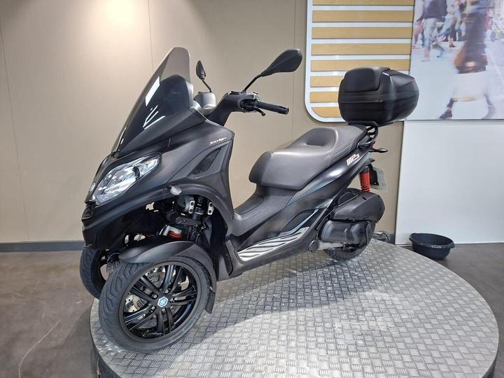 A black three-wheeled scooter with a windscreen and a rear storage box, positioned on a circular display stand in a modern showroom.