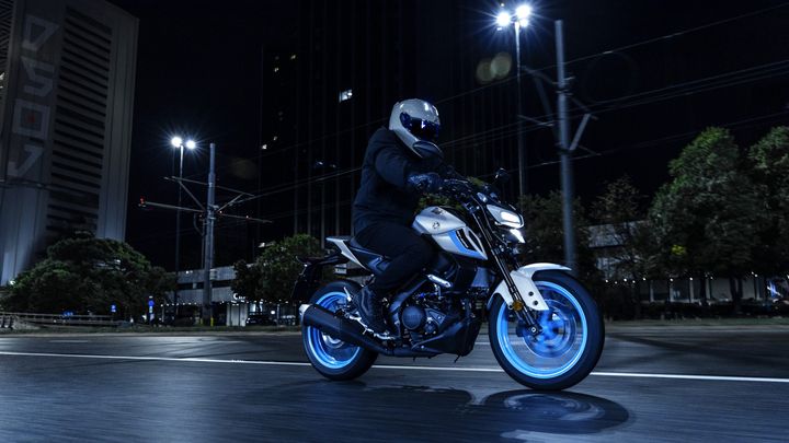 A motorcyclist wearing a helmet and dark clothing rides a sleek motorcycle with illuminated blue wheels on a city street at night, surrounded by buildings and streetlights.