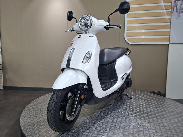 A white scooter is displayed on a circular metallic platform, showcasing its smooth body design, round headlight, and single seat. The background features a simple beige wall with a modern design element.