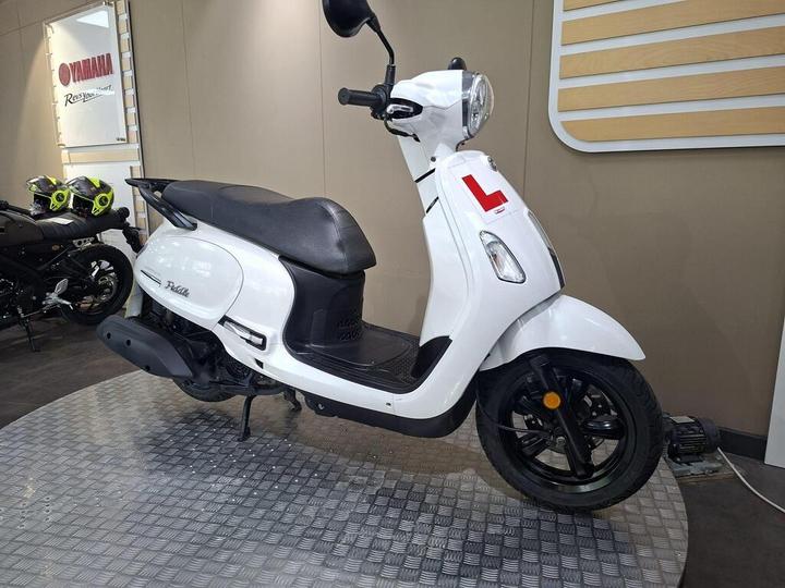 A white Yamaha scooter with a black seat displayed on a metal platform in a showroom, featuring a learner's plate and a background with Yamaha branding and motorcycle helmets.