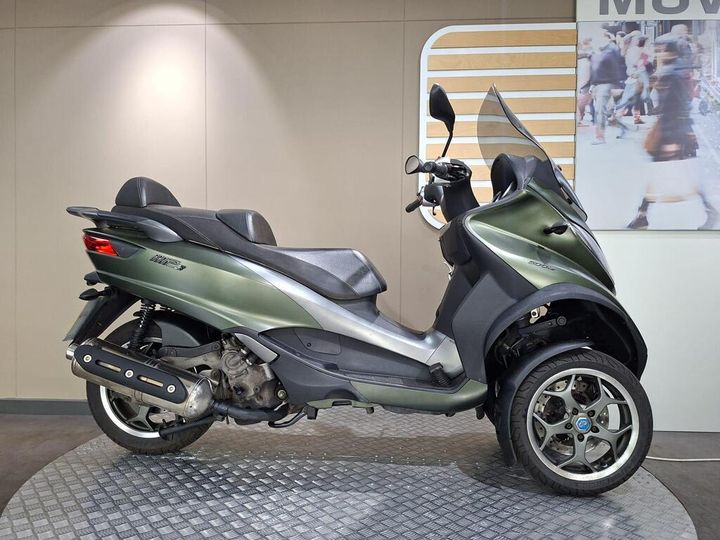 Piaggio MP3 500 LT Sport 2018 A green three-wheeled scooter positioned on a textured platform, featuring a sleek design with a double seat, a windshield, and alloy wheels. The background is a neutral wall with a faint display of pedestrians.