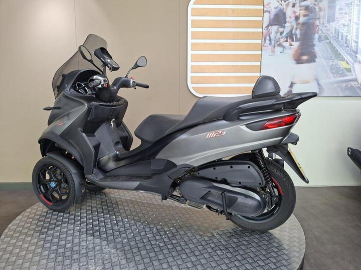 A sleek, three-wheeled scooter displayed in a showcase, featuring a dark gray body, large windshield, and prominent rear lights. The background includes a neutral-colored wall with a modern graphic of a city scene.