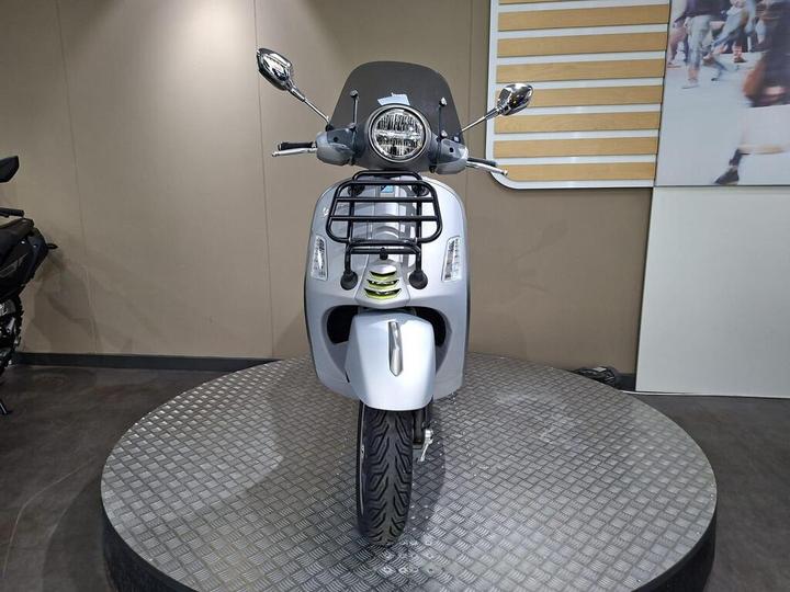 A silver scooter with a front storage rack is displayed on a round, textured platform, set against a neutral-colored wall with a wooden panel design. The scooter's mirrors and headlights are prominently visible.