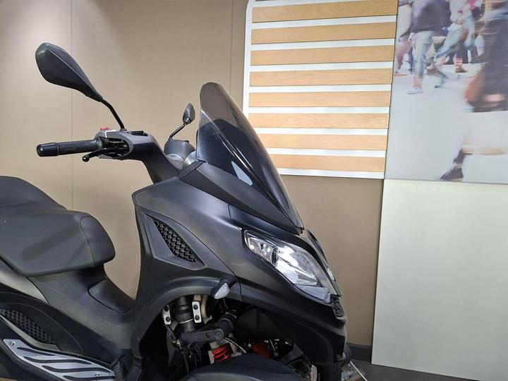 A close-up view of a black scooter with a sleek design, featuring a tinted windscreen and positioned against a beige wall with wooden slats and a blurred background of people walking.
