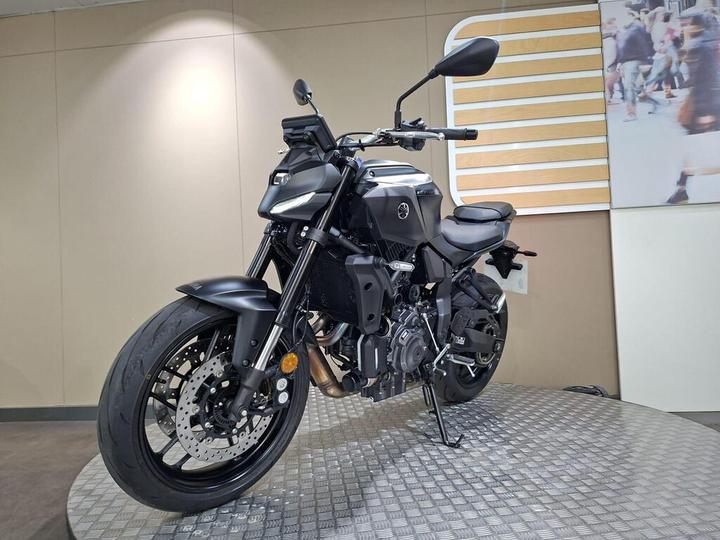 A sleek black motorcycle displayed on a textured platform, featuring a modern design with sharp lines, a prominent front headlight, and a minimalist seat, set against a neutral background with an abstract pattern.