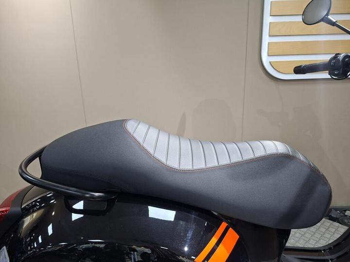 A close-up image of a motorcycle seat featuring a sleek black exterior with a gray textured section and stitching detail, mounted on a black motorcycle against a neutral background.