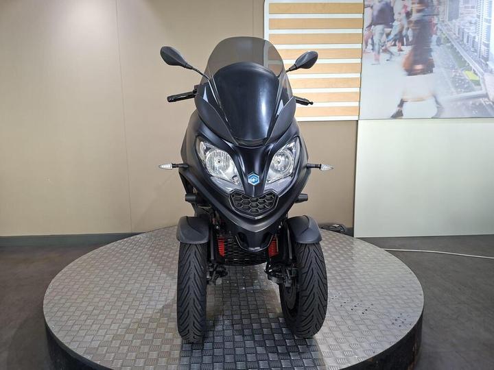 A black three-wheeled scooter is displayed front-facing on a circular platform, with a minimalistic beige and white background.