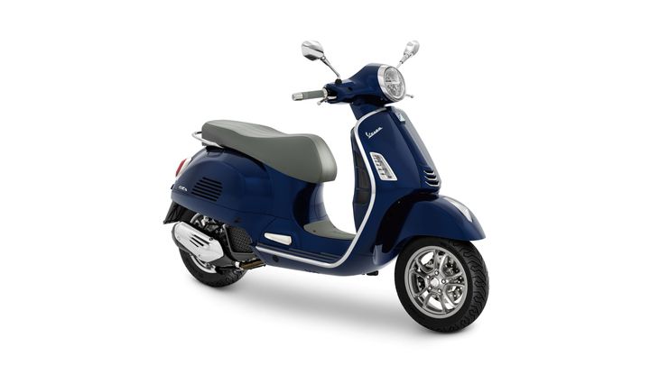 A blue Vespa scooter with a gray seat, side mirrors, and a vintage design, positioned at a slight angle against a white background.