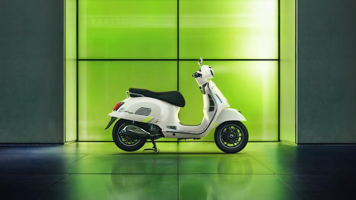 A white scooter with a black seat is positioned against a vibrant green backdrop, showcasing its sleek design and modern features. The reflection on the floor adds depth to the scene.