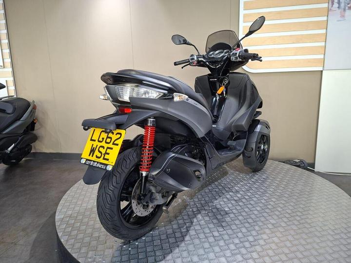 A black three-wheeled scooter is showcased on a circular display platform, featuring a red shock absorber and a registration plate labeled "LG62 WSE." The background is minimalist with light-colored walls.