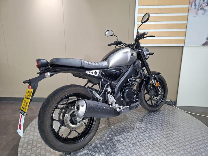 A sleek silver motorcycle is displayed on a textured platform, showcasing its modern design and black accents, with a rear view highlighting the seat and tail light.