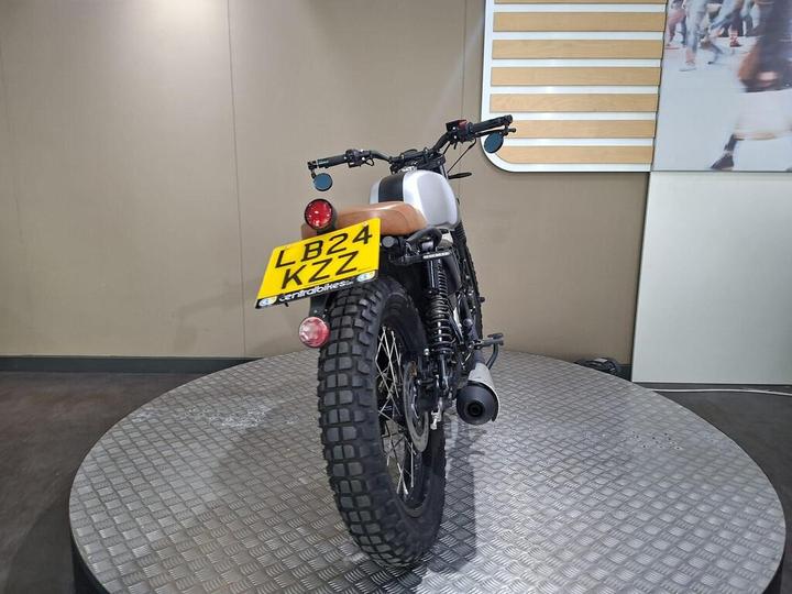 A rear view of a motorcycle on a circular display platform, featuring a black tire, a silver body with a brown leather seat, and a license plate reading "LB24 KZZ." The background is a plain wall with minimal decor.