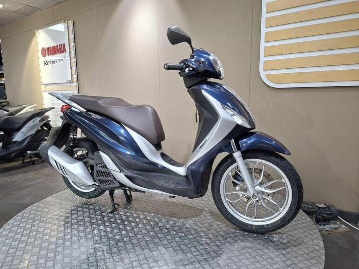 A blue and silver scooter displayed on a circular metal platform in a dealership, with a Yamaha logo visible on the wall in the background.