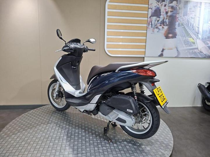 A dark blue and white scooter is displayed in a showroom, featuring a sleek design, a comfortable seat, and visible details like the rear wheel and exhaust. The background includes a light-colored wall and a large photograph of a busy city street.