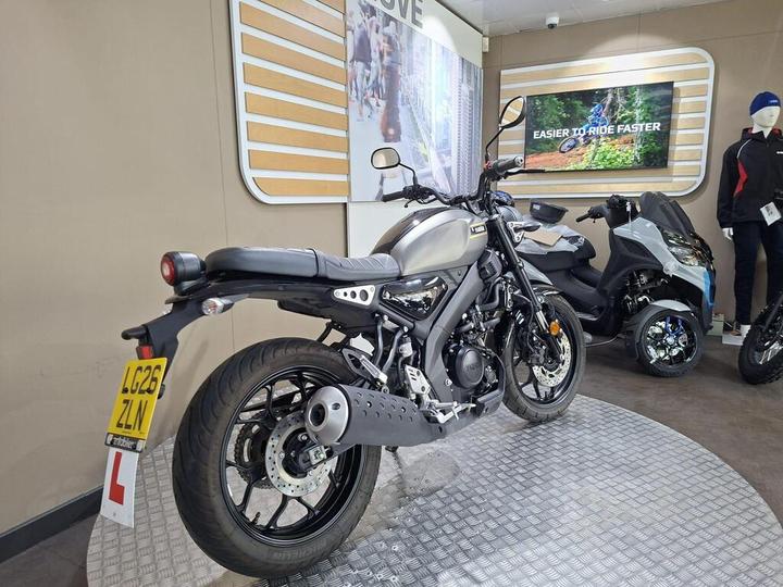 A sleek motorcycle displayed in a showroom with a circular stand, featuring a black and silver design, visible L-plate, and a backdrop of promotional graphics and a TV screen showcasing riding content. A small scooter is partially visible on the right.