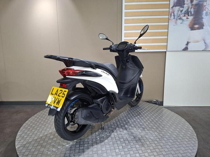 A black and white scooter displayed on a circular platform, featuring a rear view with a visible license plate, set against a neutral wall background.