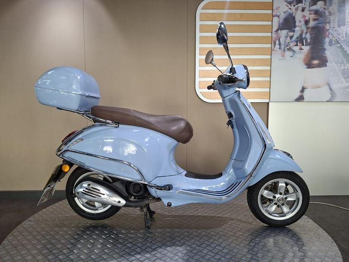 Vespa Primavera 50 2017 A blue scooter with a brown seat and a matching front storage compartment, displayed on a circular platform against a neutral background.