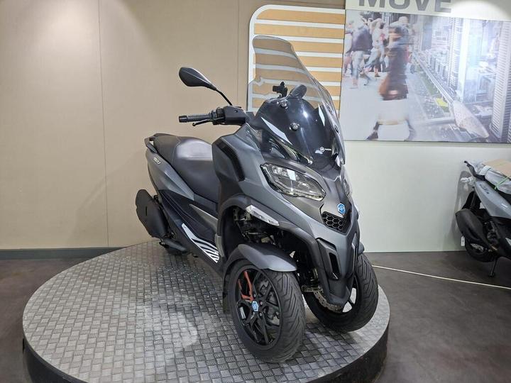 A modern gray scooter with three wheels displayed on a rotating platform against a beige wall, featuring a cityscape mural in the background.