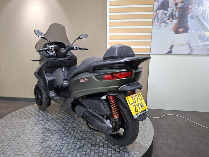 A three-wheeled scooter displayed in a showroom, featuring a sleek dark design, windshield, and a visible license plate reading "LD70 ZYM." The background includes a wall with wooden accents and a blurred image of people walking.