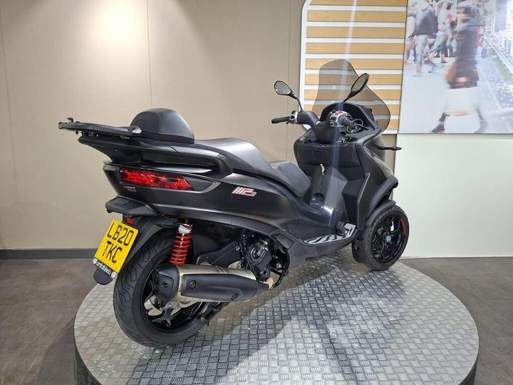 A black three-wheeled scooter displayed on a checkered platform, with a transparent windscreen and rearview mirrors, against a backdrop of a modern indoor space. The scooter has a visible license plate, and its design features sporty lines and red shock absorbers.