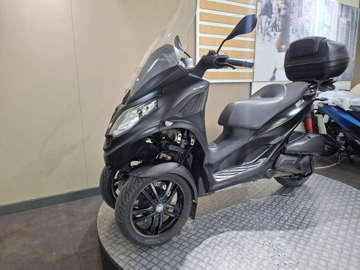 A black three-wheeled scooter with a windshield, featuring a sporty design and a storage compartment on the rear, displayed on a textured circular platform in a showroom.