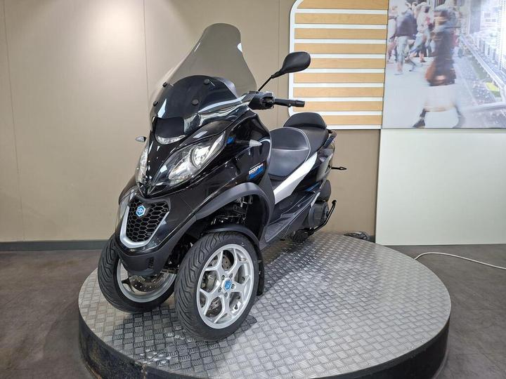 A black three-wheeled scooter on a circular display platform, featuring a windscreen and modern design elements. The background includes a light-colored wall and a blurred image of pedestrians.