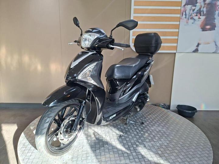 A black scooter displayed on a circular platform with a textured surface, featuring a windshield, rear-view mirrors, and a storage compartment on the back. The background has a simple, neutral wall and a blurred image of people.