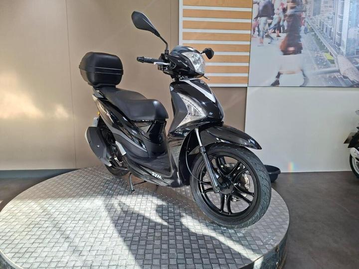 A black scooter displayed on a circular platform with a textured surface, featuring a rear storage box, set against a light-colored wall and a backdrop of an urban scene.