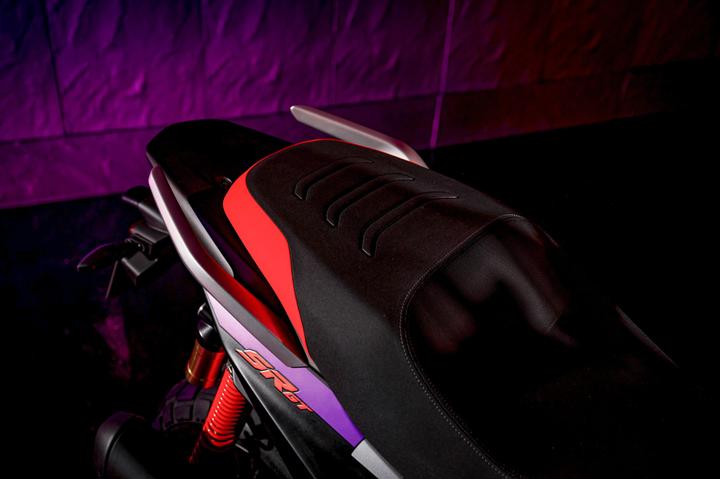 A close-up of a motorcycle seat featuring a sleek design with black material highlighted by red accents, set against a gradient purple and black background. The motorcycle's brand logo is subtly visible on the side.