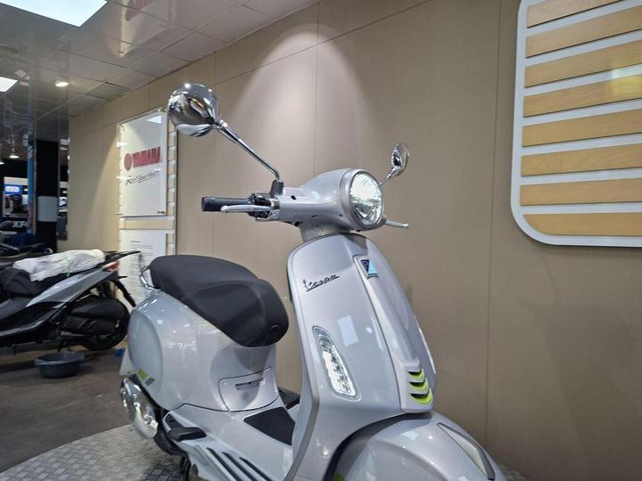 A silver scooter with a rounded front, featuring a prominent headlight and a rearview mirror, is displayed against a neutral-colored wall with a Yamaha logo and signage in the background.